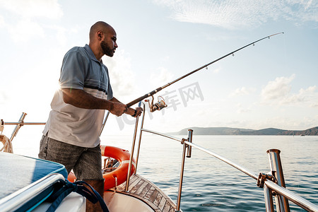 日落时，年轻的非洲裔美国男子拿着钓鱼竿站在帆船上钓鱼
