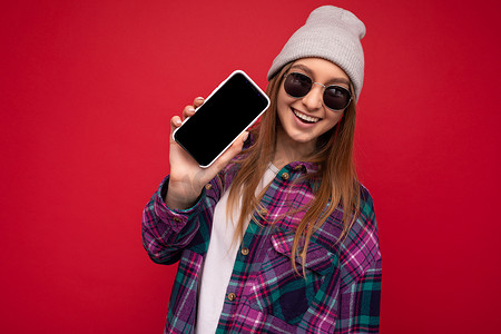 照片中，美丽而积极的年轻金发女子穿着时尚的紫色衬衫和休闲的白色 T 恤灰色帽子和太阳镜，在红色背景上被隔离，手拿手机，屏幕上有空屏幕，用于观看相机的模型