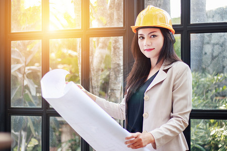 工业文件摄影照片_美丽的亚洲女建筑师的肖像，戴着黄色头盔安全帽研究房间的蓝图计划，严肃的土木工程师在建筑工地处理文件