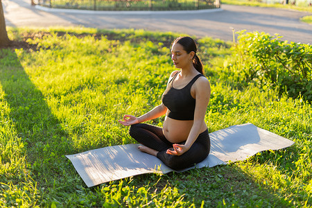 在阳光明媚的温暖夏日，身着体操服的平静的年轻积极孕妇做瑜伽，坐在绿草上的垫子上冥想。
