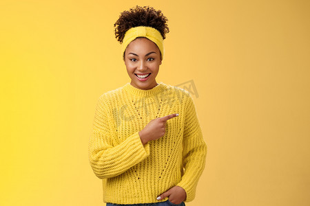 黄色促销摄影照片_友好的、轻松的非洲裔美国女性在谈话中随意指着右边，讨论最近新开的咖啡馆开张的折扣促销，愉快地站在黄色背景上