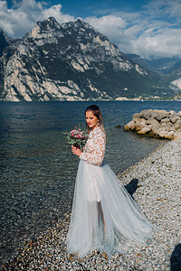 加衣服摄影照片_一个穿着漂亮白色连衣裙的女孩沿着加尔达湖的堤岸行走。一名妇女在意大利的山和湖的背景下被拍照。托尔博莱