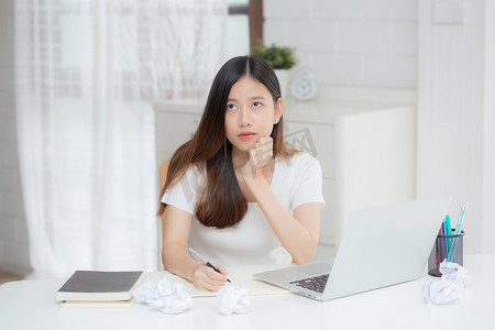 使用笔记本电脑的年轻亚洲女性认为创意项目和纸张在家里的桌子上出现问题，女孩使用笔记本电脑时感到沮丧和麻烦、商业和自由职业概念。