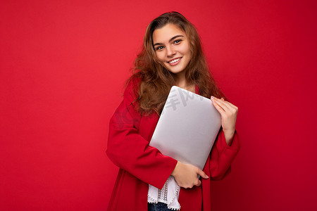 照片中，美丽微笑、快乐的黑发年轻女子手持电脑笔记本电脑，身穿红色羊毛衫和白色衬衫，看着红色背景中突显的相机