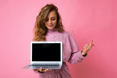 照片中，美丽的金发卷发年轻女子看着上网本键盘，手里拿着电脑笔记本电脑，笔记本电脑上有空的显示器屏幕，上面有模型和复制空间，一根手指穿着粉色连帽衫，与粉色墙壁背景隔离