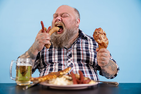 饥饿的大号男人在餐桌上吃香肠，配上丰富的食物和一杯啤酒