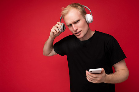 心烦意乱地询问穿着黑色 T 恤和白色耳机的英俊金发年轻人站在红色背景上，拿着智能手机和在线交流听音乐并享受相机的乐趣