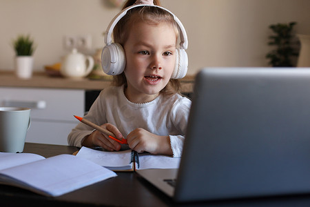 戴着耳机的微笑小女孩在家中使用笔记本电脑在线手写学习，戴着耳机的可爱快乐小孩子在电脑上上互联网网络课程或课程。