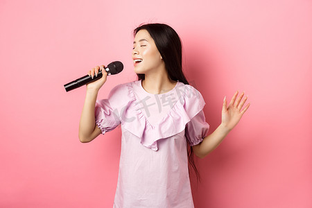 站立的麦克风摄影照片_美丽的亚洲女孩表演歌曲，在麦克风里唱歌，浪漫地微笑着，穿着粉红色背景的裙子站立