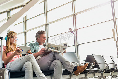年轻漂亮的少女戴着耳机在数字平板电脑上看电影，而她的父亲正在机场看报纸