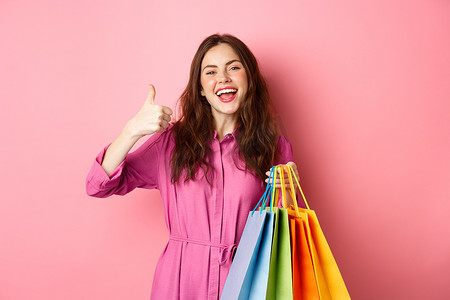 年轻快乐的女购物者竖起大拇指，对优惠折扣感到满意，购买人员打折，拿着购物袋，微笑着高兴，粉红色的背景