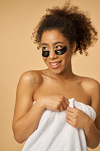 淋浴后裹着白毛巾的年轻女子看起来很顽皮，微笑着，在米色背景中隔离的眼罩下涂着黑色的姿势