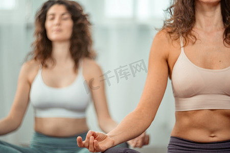 两名身穿运动上衣和紧身裤的年轻健康女性在室内练习瑜伽，坐在瑜伽垫上的莲花姿势上，闭着眼睛冥想放松。