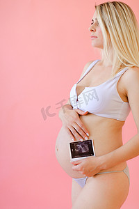 年轻怀孕女孩抱着腹部在粉红色单声道背景下与超声波。