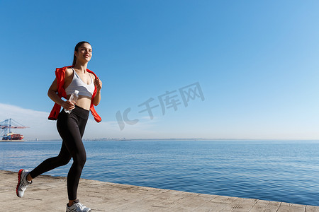 跑步风景摄影照片_微笑的女运动员沿着海边跑步并欣赏风景的户外照片。
