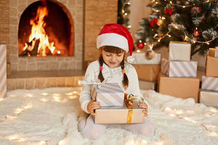 平安夜，壁炉旁快乐的小孩子坐在柔软的地毯上，手里拿着两个礼物盒，表情快乐地看着她的礼物，想要打开它们，穿着休闲服，戴着帽子