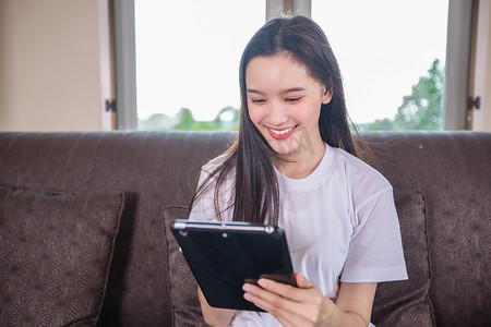 正在玩电脑的人摄影照片_亚洲年轻女性快乐开朗可爱美丽正在玩数字平板电脑，坐在家里的沙发上，微笑的女士使用应用阅读电子书在客厅放松