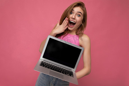 美丽而惊讶的年轻女子拿着电脑笔记本电脑，显示器上空着，穿着粉色作物上衣，看着粉色背景中突显的相机
