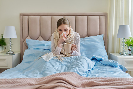 家里生病的年轻女子躺在床上，手里拿着一杯热饮和手帕