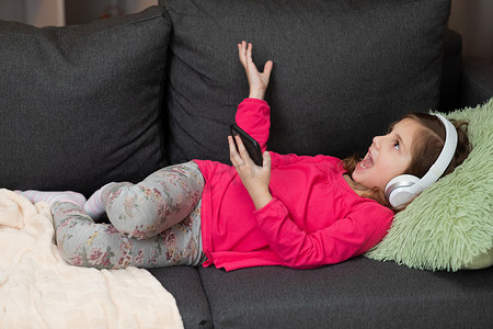 戴着无线耳机的小女孩在沙发上冷笑，拿着手机使用移动在线播放器应用程序听最喜欢的音乐。