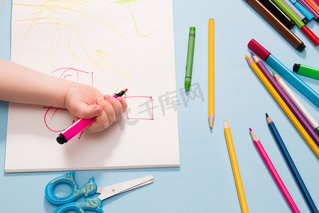 一个小孩用铅笔在相册、复印空间、顶视图、蓝色背景、铅笔、蜡笔、剪刀和孩子的手在桌子上画画