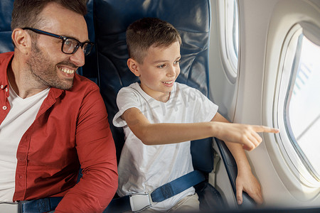 快乐的小男孩看着窗外，坐在窗边向父亲展示一些东西，一起乘飞机旅行