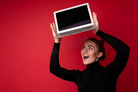 职业照摄影照片_照片中，美丽微笑、快乐的黑发年轻女子拿着电脑笔记本电脑，显示器屏幕空空如也，穿着黑色长袖，抬头看着红色背景中突显的上网本