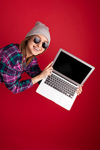 疯狂疯狂欣喜若狂的特写肖像美丽微笑快乐的年轻女子拿着上网本电脑看着相机享受戴着太阳镜彩色衬衫灰色帽子隔离在红墙背景