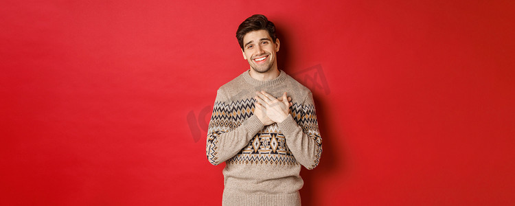 感动而快乐的帅哥肖像，收到新年礼物，手握心微笑，说谢谢，站在红色背景下的圣诞毛衣