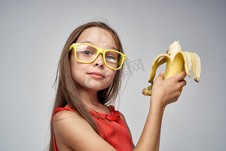 连衣裙戴眼镜背包摄影照片_戴眼镜的小女孩，穿着红色连衣裙，手里拿着香蕉，浅色背景