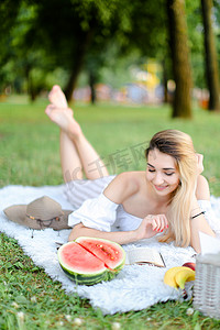 年轻漂亮的女人躺在公园的格子上，在西瓜和帽子旁边看书。