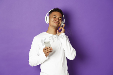 无忧无虑的非洲裔美国人的形象，享受着耳机听音乐，拿着智能手机，高兴地微笑，站在紫色背景上
