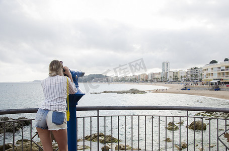 在阳光明媚的日子里，在西班牙布拉瓦海岸 Lloret de Mar 的海滩上，穿着白衬衫和带着黄色小包的迷人年轻女子用双筒望远镜望向大海。