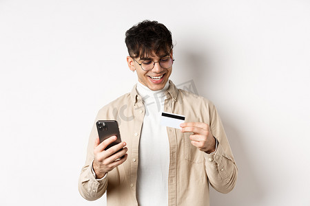 戴眼镜的快乐年轻人在网上购物，看着塑料信用卡，拿着智能手机，站在白色背景上