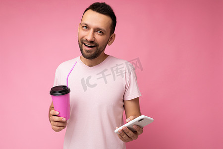 积极微笑的英俊的年轻不刮胡子的黑发男子，留着胡子，穿着日常浅粉色 T 恤，在粉色背景下被隔离，拿着和使用手机上网冲浪，看着相机喝饮料