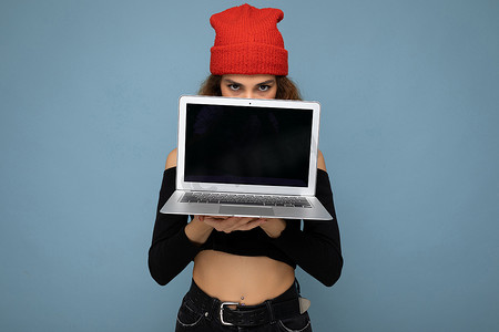 滑稽美丽的黑金发女人的特写肖像，她拿着笔记本电脑，看着相机，身穿黑色作物上衣，红色和橙色的抹布隔离在浅蓝色的墙壁背景上