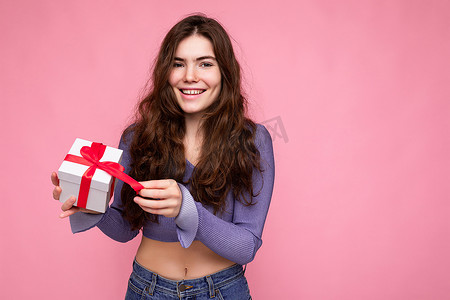 拆圣诞节礼物摄影照片_一张迷人的、积极微笑的黑发卷发年轻女子的照片，被隔离在粉红色背景墙上，穿着紫色衬衫，拿着白色礼盒，上面有红丝带，拆箱礼物看着相机