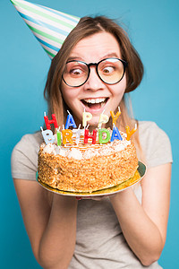 节日蓝色海报摄影照片_戴着纸帽子和眼镜的疯狂有趣的女孩，在蓝色背景上拿着一个大生日蛋糕。