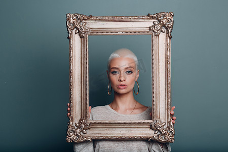千禧年摄影照片_金发短发的千禧年年轻女子手里拿着镀金相框，背后是她的脸部肖像。