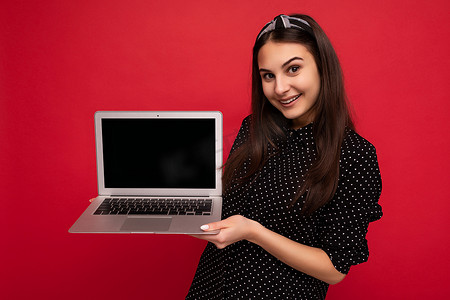 照片中，身穿黑色衣服的美丽微笑深色女孩拿着电脑笔记本电脑，看着彩色墙壁背景上突显的相机