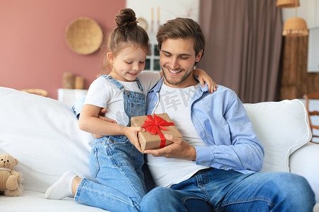 礼盒惊喜摄影照片_可爱的小女儿给爸爸带来惊喜，小女孩向坐在沙发上的父亲赠送礼盒。