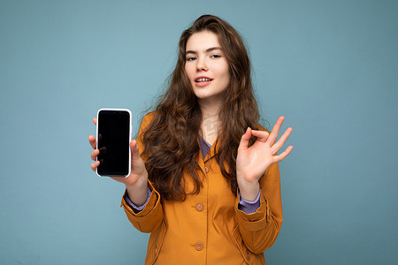 美丽的黑发年轻女子穿着橙色夹克，在蓝色背景中被隔离，手拿手机，展示着空显示屏的手机，用于模型看着相机，并表现出良好的手势