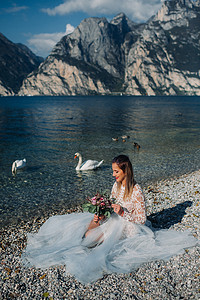 一个穿着漂亮白色连衣裙的女孩坐在加尔达湖的堤岸上。在意大利的山和湖的背景下拍摄了一个女人的照片。托尔博莱