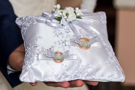 结婚金色摄影照片_新郎拿着一个带结婚戒指的白色枕头