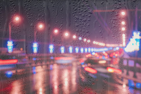 行驶中的汽车和灯笼的模糊灯光反射在夜城被雨覆盖的窗户后面潮湿的沥青上。