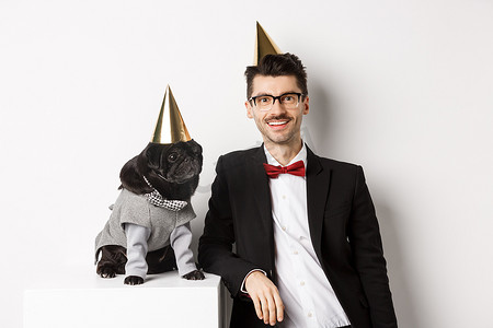 英俊的年轻人与穿着派对服装、头戴圆锥体的可爱黑色哈巴狗庆祝生日的照片，站在白色背景上