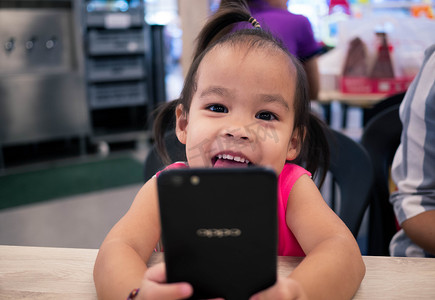 在购物中心的美食广场，亚洲小孩正在智能手机上愉快地看卡通片。