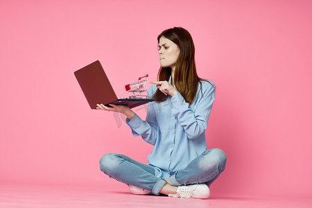 购物娱乐摄影照片_坐在地板上、带笔记本电脑购物娱乐粉红色背景的女人