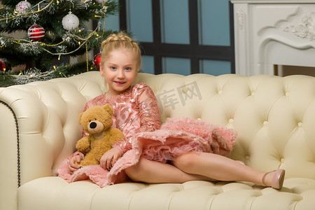 一个抱着泰迪熊的小女孩坐在圣诞树旁的沙发上。