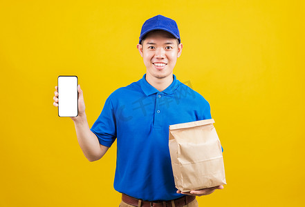 手机容器摄影照片_穿着蓝色制服微笑的送货服务男子拿着纸容器，用于外卖袋杂货食品包并展示智能手机